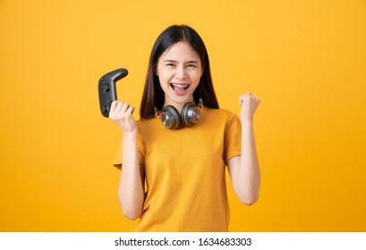 Cheerful Beautiful Asian Woman In Casual Yellow T-shirt And Playing Video Games Using Joysticks With Headphones On Orange Background.