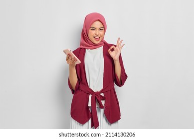 Cheerful beautiful Asian woman in casual shirt and hijab holding mobile phone and gesturing ok sign isolated over white background. People religious lifestyle concept - Powered by Shutterstock