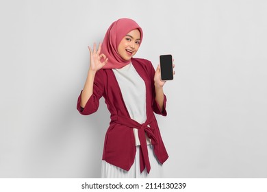 Cheerful beautiful Asian woman in casual shirt and hijab holding blank mobile phone screen, showing ok sign isolated over white background. People religious lifestyle concept - Powered by Shutterstock
