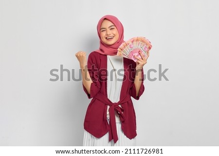 Cheerful beautiful Asian woman in a casual shirt and hijab showing Indonesian rupiah banknotes, celebrating luck isolated over white background. People religious lifestyle concept