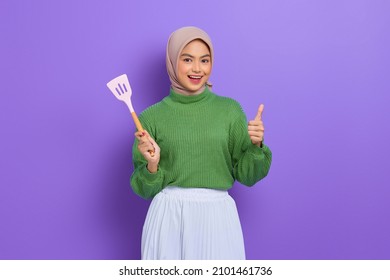 Cheerful beautiful Asian woman in green sweater and hijab holding a spatula and showing thumb up isolated over purple background - Powered by Shutterstock