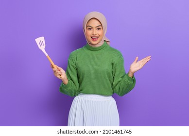 Cheerful beautiful Asian woman in green sweater and hijab holding spatula isolated over purple background - Powered by Shutterstock