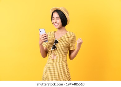 Cheerful beautiful Asian woman holding smartphone with fists clenched celebrating victory expressing success on light yellow background.