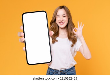Cheerful beautiful Asian woman holding smartphone and shows ok sign on light yellow background.