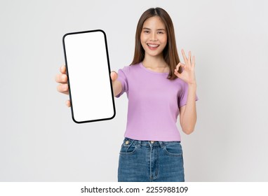 Cheerful beautiful Asian woman holding big smartphone and shows ok sign on grey background.