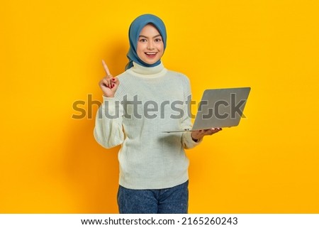 Cheerful beautiful Asian woman in white sweater using laptop and pointing fingers up having a good idea isolated over yellow background. people religious lifestyle concept