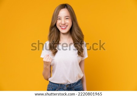 Cheerful beautiful Asian woman in a white t-shirt raises arms and fists clenched with shows strong powerful, celebrating victory expressing success.