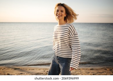 Cheerful Beautiful Caucasian Young Woman Smiling With Teeth Looking Into Camera On Background Of Sea. Her Blonde Short Hair Blowing In Wind, Wearing Striped Blouse And Jeans.