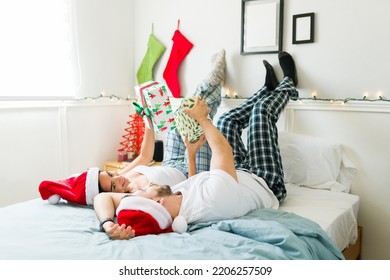 Cheerful Beautiful Couple Exchanging Christmas Presents With Their Feet Up In Bed While Relaxing Together In The Bedroom