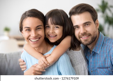 Cheerful beautiful family of three embracing laughing looking at camera, happy cute kid girl hugging smiling mom posing with dad for portrait, child daughter mother and father bonding headshot