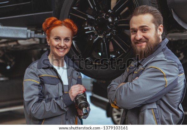 Cheerful beautiful female mechanic and her male\
colleague smiling to the camera, working at car service station.\
Experienced car technicians repairing vehicles at the garage.\
Female equality\
concept