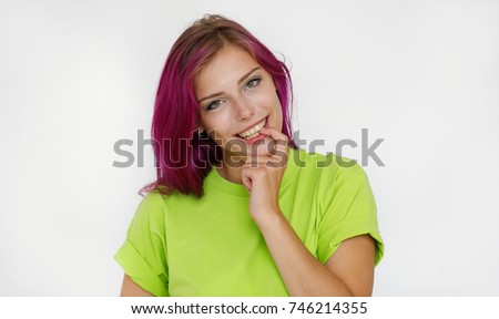 Cheerful beautiful girl with purple hair dressed in a green t-shirt looks with a smile at the camera biting one finger with teeth against a white wall background. Healthy lifestyle concept.
