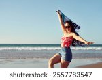 cheerful beautiful latina woman walking holding a towel upwind on the beach, summer and vacation concept
