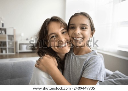 Cheerful beautiful mom and happy tween daughter kid hugging with love, faces touch, looking at camera, smiling, showing white teeth, posing for home close up portrait, talking on video call