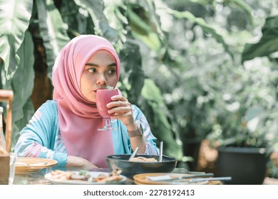 Cheerful and beautiful Muslim Asian woman in a pink hijab drinking a berry fruit smoothie at an outdoor restaurant - Powered by Shutterstock