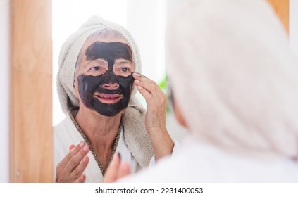 Cheerful beautiful senior woman in bathrobe applying a detox facial charcoal mask homemade looking at the mirror - wellness, take care of the skin concept - Shutterstock ID 2231400053