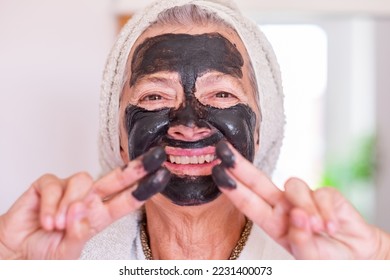 Cheerful beautiful senior woman in bathrobe applying a detox facial charcoal mask homemade - wellness, take care of the skin concept - Shutterstock ID 2231400073