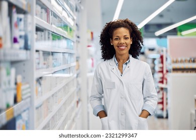 A cheerful beautiful smiling African-American female pharmacist with curly hair, standing next to the pharmacy shelves. - Powered by Shutterstock