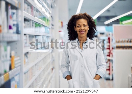A cheerful beautiful smiling African-American female pharmacist with curly hair, standing next to the pharmacy shelves.