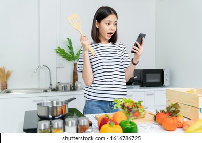 Cheerful beautiful woman hand holding a smartphone and salad bowl with ladle and various green leafy vegetables on the table. Talking on the mobile with a friend in the morning in the kitchen - Powered by Shutterstock