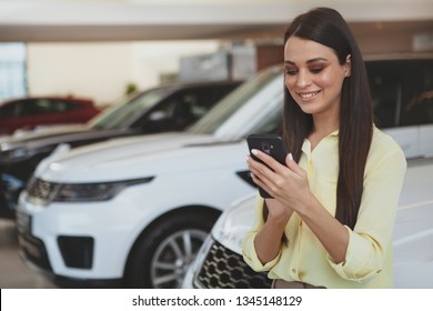 Cheerful Beautiful Woman Smiling, Typing Messages On Her Smart Phone While Shopping For A New Car At The Dealership Salon. Attractive Female Customer Buying New Automobile