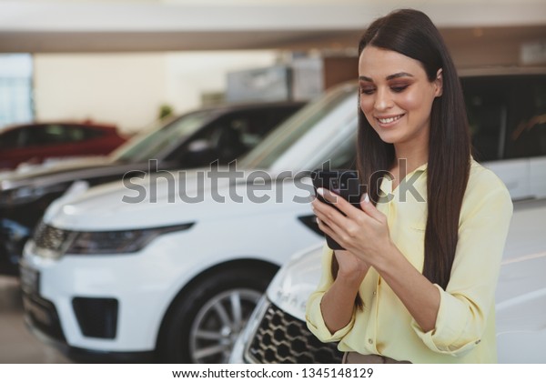 Cheerful beautiful woman\
smiling, typing messages on her smart phone while shopping for a\
new car at the dealership salon. Attractive female customer buying\
new automobile