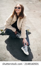 Cheerful Beautiful Woman In Trendy Clothes Having Fun While Sitting On Skateboard Outdoors. Urban Area With Modern Building On Background. Lifestyles Of Youth People.