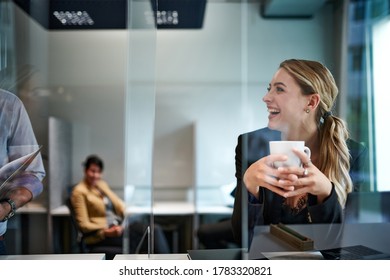 Cheerful Beautiful Young Blond Businesswoman Holding Cup While Looking At Colleague In Office Seen Through Glass Shield