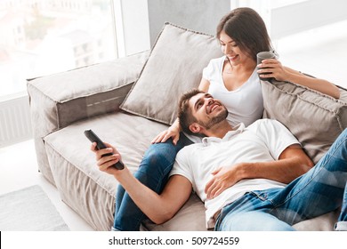 Cheerful beautiful young couple drinking coffee and watching TV at home