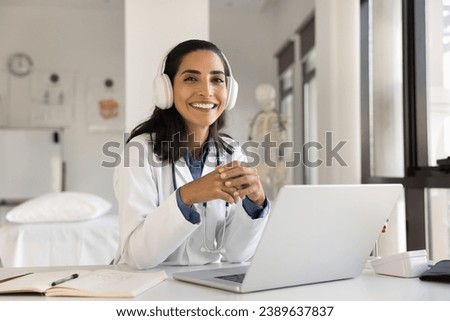 Cheerful beautiful young doctor woman in head phones posing at hospital workplace with laptop computer, looking at camera with toothy smile, offering online medical consultation. Professional portrait