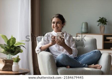 Cheerful beautiful young Indian woman holding cell, sitting in armchair at home, looking away, smiling, laughing, chatting online, typing, ysing app on Internet, thinking over text message
