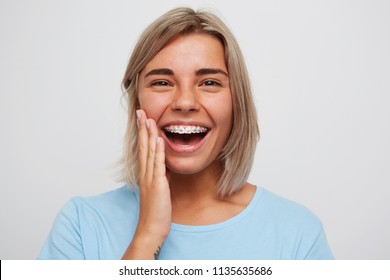 Cheerful beautiful young woman with blonde hair and braces on teeth laughing and touching her face by hand isolated over white background - Shutterstock ID 1135635686