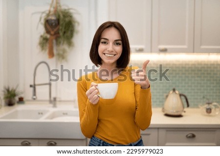 Cheerful beautiful young woman enjoying herbal tea at home, standing at modern cozy kitchen, holding coffee mug and showing thumb up at camera, woman relaxing after working day, copy space