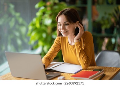 Cheerful beautiuful brunette young woman in smart casual entrepreneur attending online business meeting, woman sit at table in front of laptop, using earpods, taking notes, cafe interior, copy space