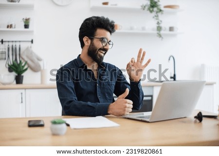 Cheerful bespectacled adult waving hello at webcam of portable computer while greeting colleague in home office. Proficient indian consultant receiving online conference call using technologies.