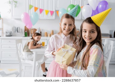 cheerful birthday girl in party cap receiving present from friend while looking together in camera - Powered by Shutterstock