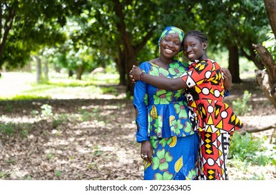 Cheerful black African teenager hugging her mother both standing in a forest among mango trees; mother-daughter relationship concept