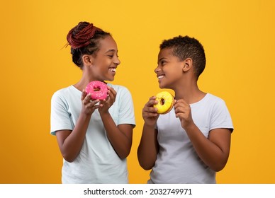 Cheerful black boy and girl siblings eating delicious donuts, looking at each other and smiling, happy afro-american brother and sister holding tasty sweets, yellow studio background