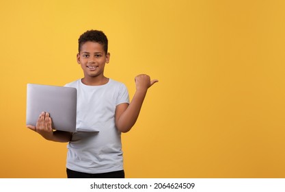 Cheerful black boy schooler with laptop posing on yellow studio background, pointing at copy space for advertisement, panorama. African american kid playing video games on computer