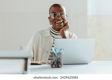 Cheerful black businesswoman with glasses working on a laptop at a bright modern office, exuding positivity and job satisfaction. Casual professional, creative workspace, happy employee. - Powered by Shutterstock