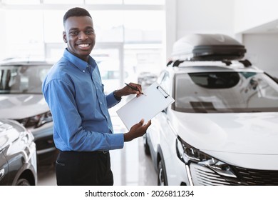 Cheerful Black Car Salesman Holding Empty Clipboard, Smiling At Camera In Modern Auto Dealership, Mockup. Positive Sales Agent Selling Luxury Vehicles At Showroom Store, Copy Space