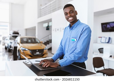 Cheerful Black Car Salesman Posing At Work Desk, Smiling At Camera, Typing On Laptop In Auto Dealership. Happy African American Automobile Manager In Showroom Store, Copy Space