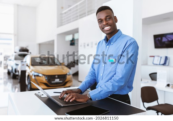 Cheerful black car
salesman posing at work desk, smiling at camera, typing on laptop
in auto dealership. Happy African American automobile manager in
showroom store, copy
space