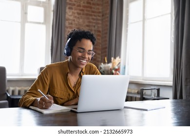 Cheerful Black college student in earphones attending virtual class, writing notes, watching webinar on Internet, studying online. African worker in headphones talking to customer on video call