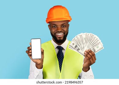 Cheerful Black Construction Worker Holding Money Cash And Blank Smartphone While Posing Over Blue Background, Happy Man In Hard Hat And Safety West Recommending App With Online Profit, Mockup