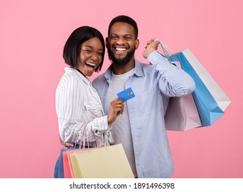 Cheerful black couple with credit card and shopping bags, buying gifts for Valentine's Day remotely on pink background. Positive young guy and his girlfriend encouraging contactless payment