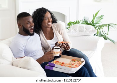 Cheerful Black Couple Eating Pizza And Drinking Coffee, Sitting On Couch At Home, Enjoying Weekend Together.