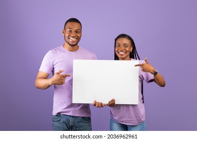 Cheerful Black Couple Holding Blank Paper Poster Together And Pointing At Free Space For Your Text Smiling To Camera Standing Over Purple Violet Studio Background. People With White Board For Advert