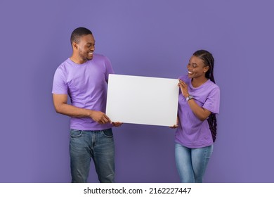 Cheerful Black Couple Holding And Showing Blank Paper Poster For Your Text Smiling Looking At Placard Standing Isolated On Purple Violet Studio Wall. Spouses With White Square Board For Advertising