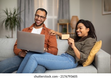 Cheerful Black Couple Shopping Online Using Laptop Computer And Credit Card Making Payment Sitting On Sofa At Home. Spouses Paying Buying Something. Internet Banking And Commerce Concept